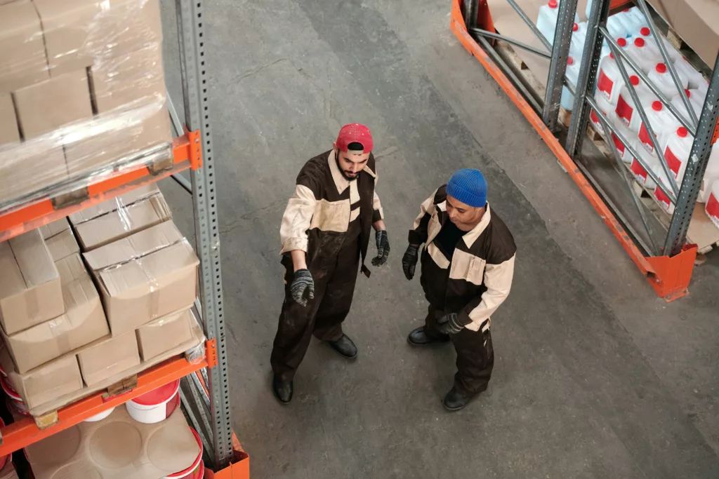 Two adult men in a warehouse communicating about storage logistics, surrounded by shelves of packages.